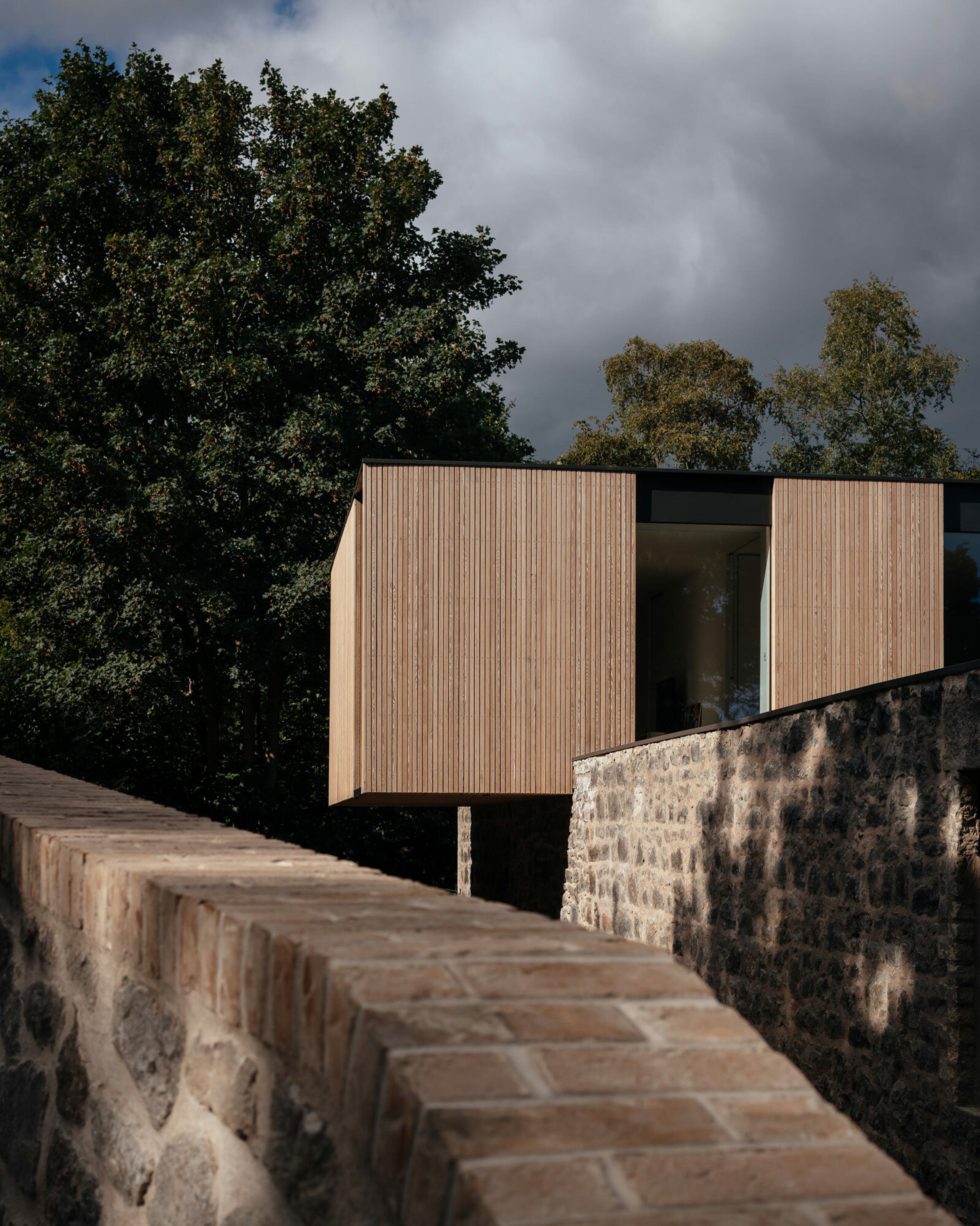 A cantilevered, timber clad house. A granite stone boundary wall surrounds the property.
