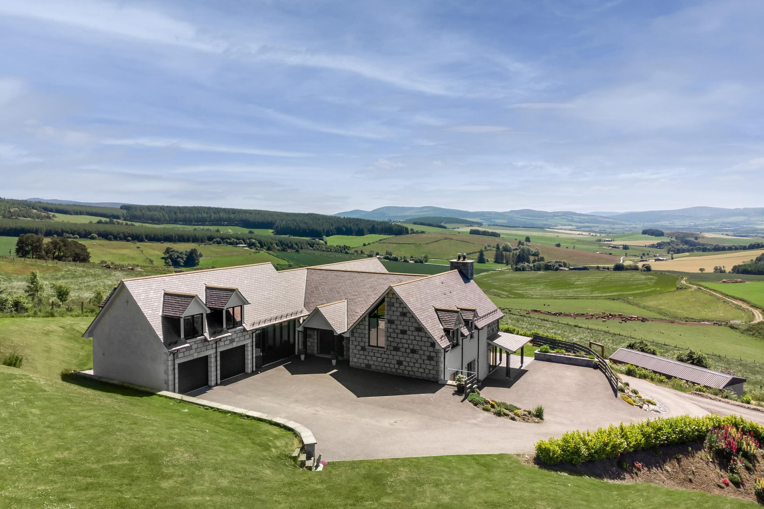 Exterior image of a large, grey granite home with a slate roof and double car garage, surrounded by green grass and farmer’s fields.