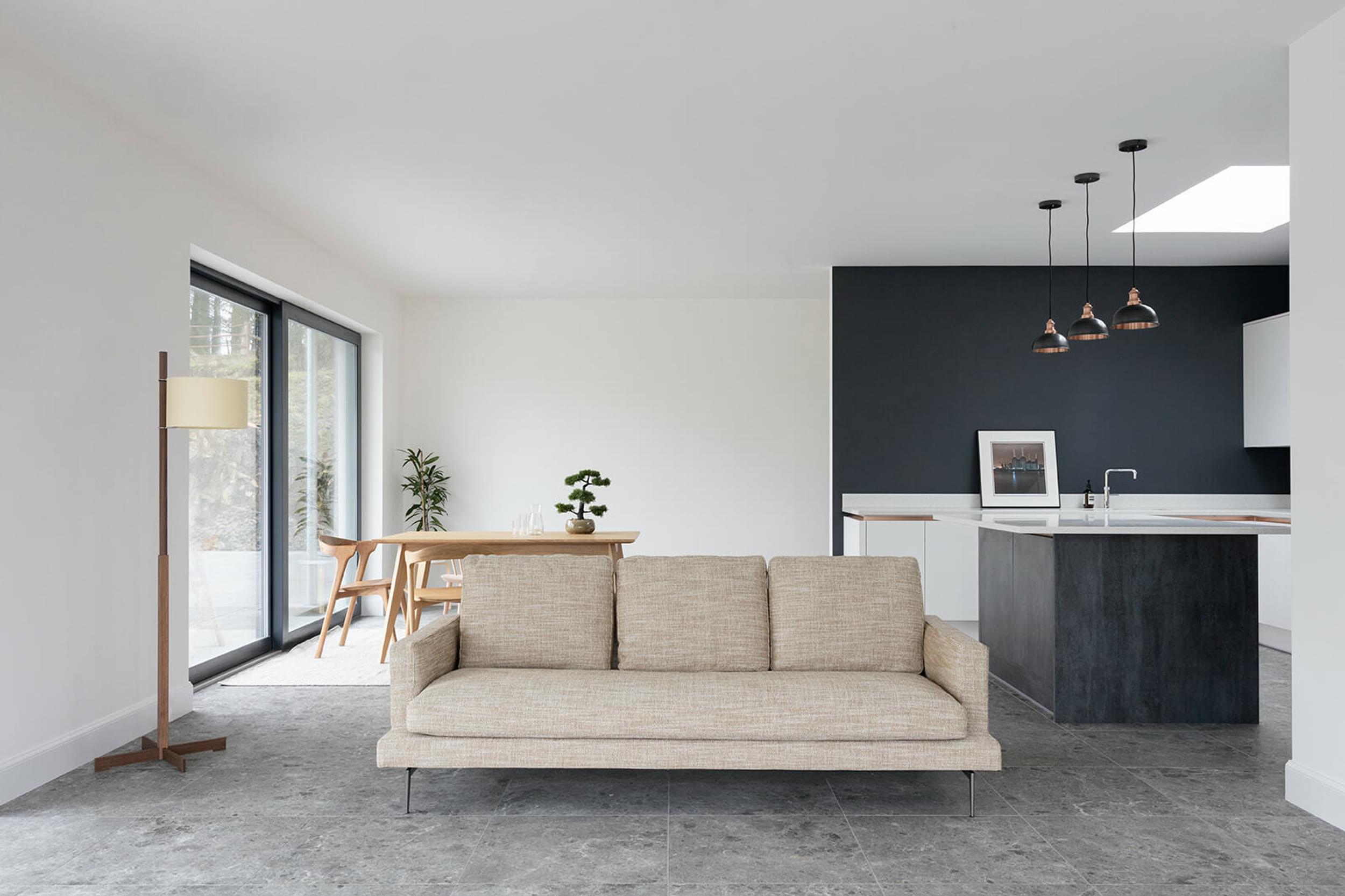 A living room setting. With a couch in the foreground and a wooden dining table in the background. A lamp stands in the room. A kitchen island is behind the couch. The walls are painted black and white.