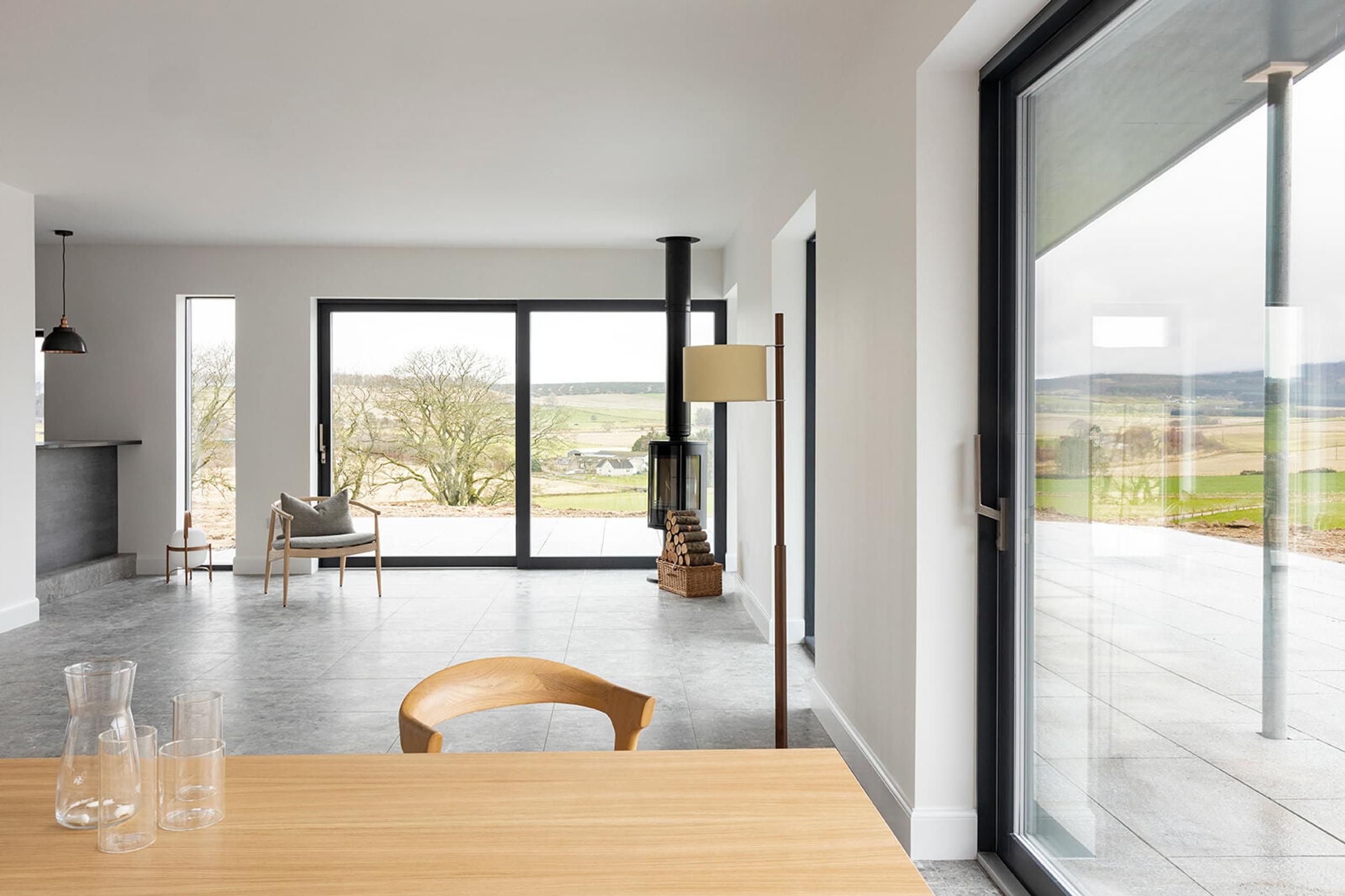 A living room with a panoramic rural view, a dining table and a chair. A black wood burning stove in the corner of the room with logs next to it. A glass of water sits on top of the table.