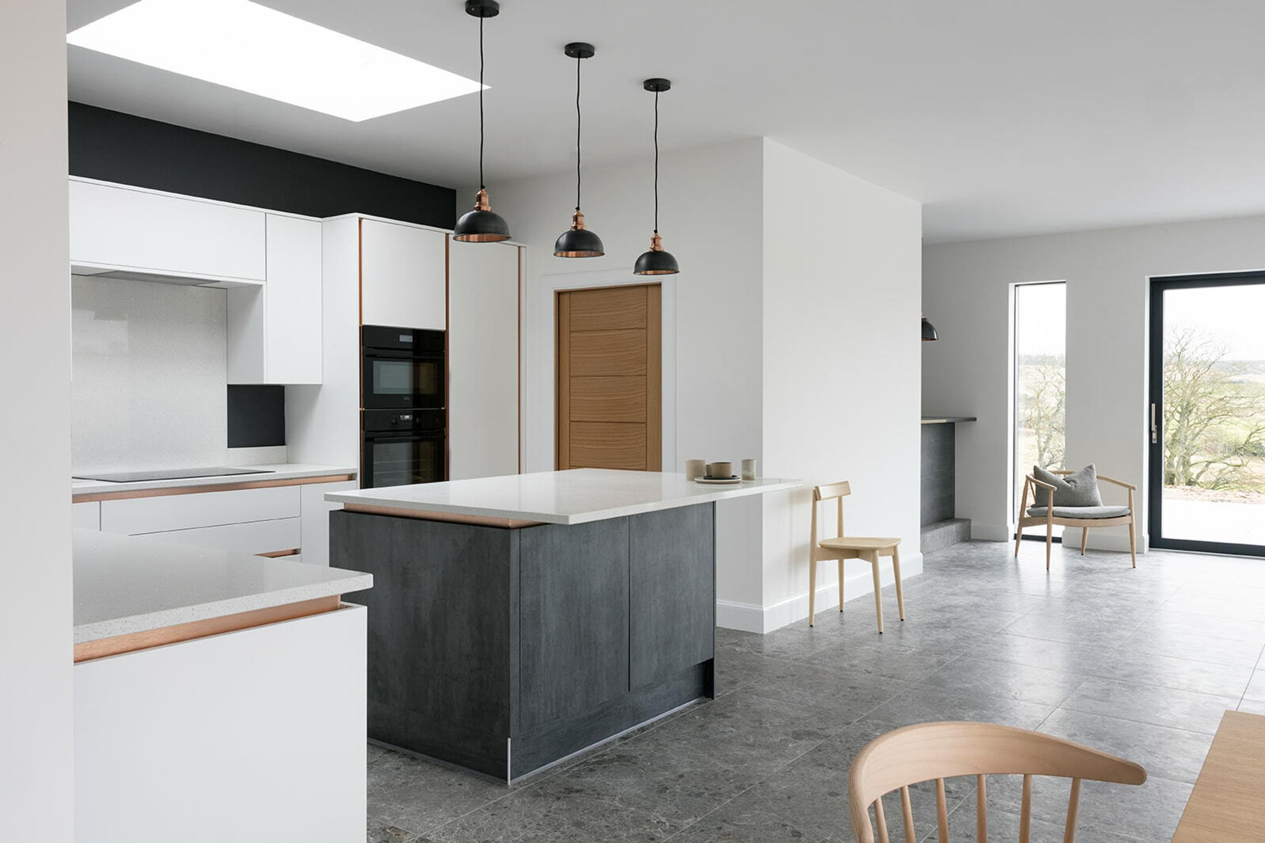 A view into a white kitchen. Three black and copper lamps hang over the black and white kitchen island. Tiles are grey. A chair sits in the corner of the room.