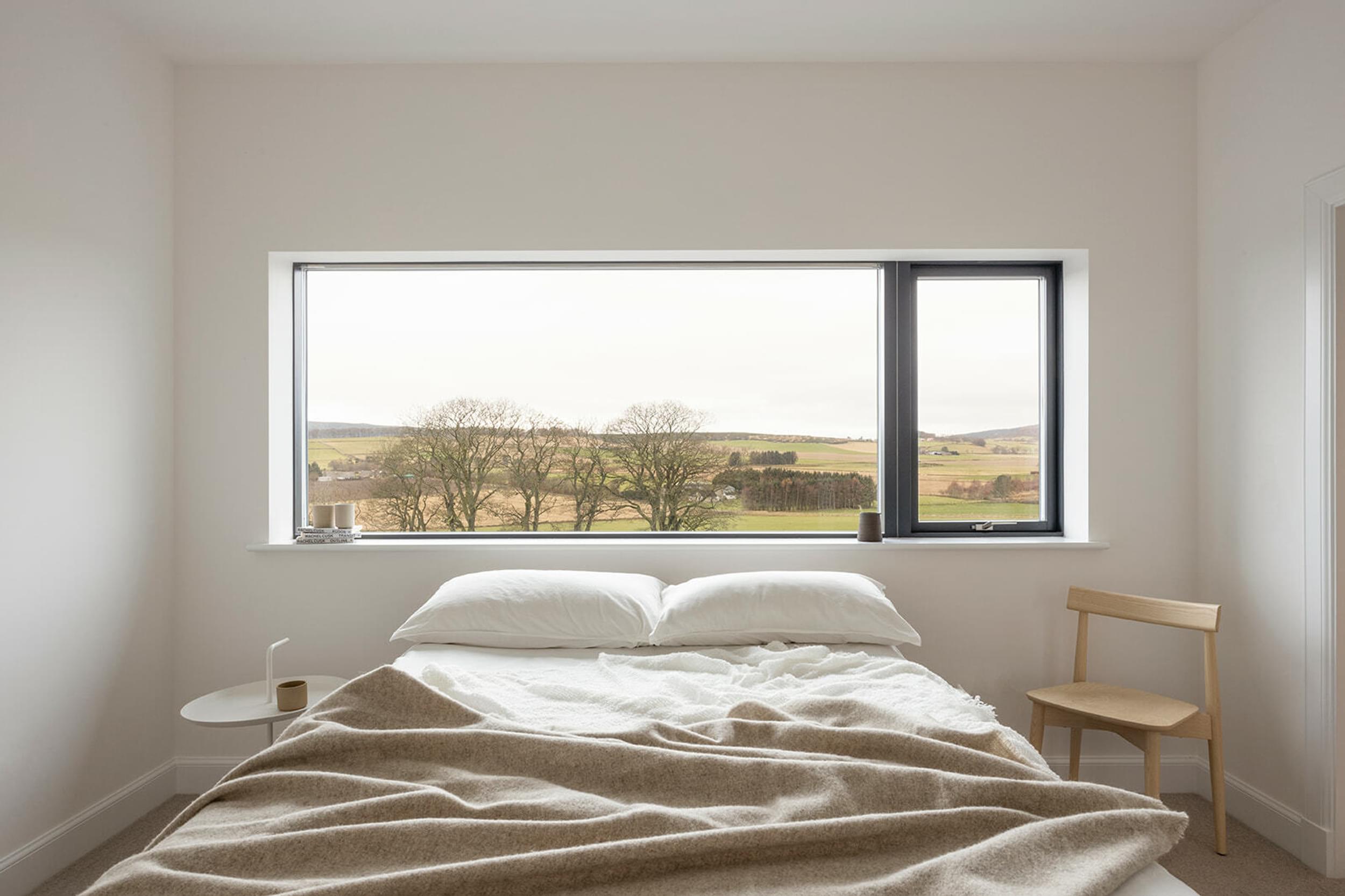 A white bedroom with a bed, white sheets and pillows and a beige throw at the foot of the bed. A long, rectangle window sits above the bed head, revealing bare trees outside and a rural landscape.
