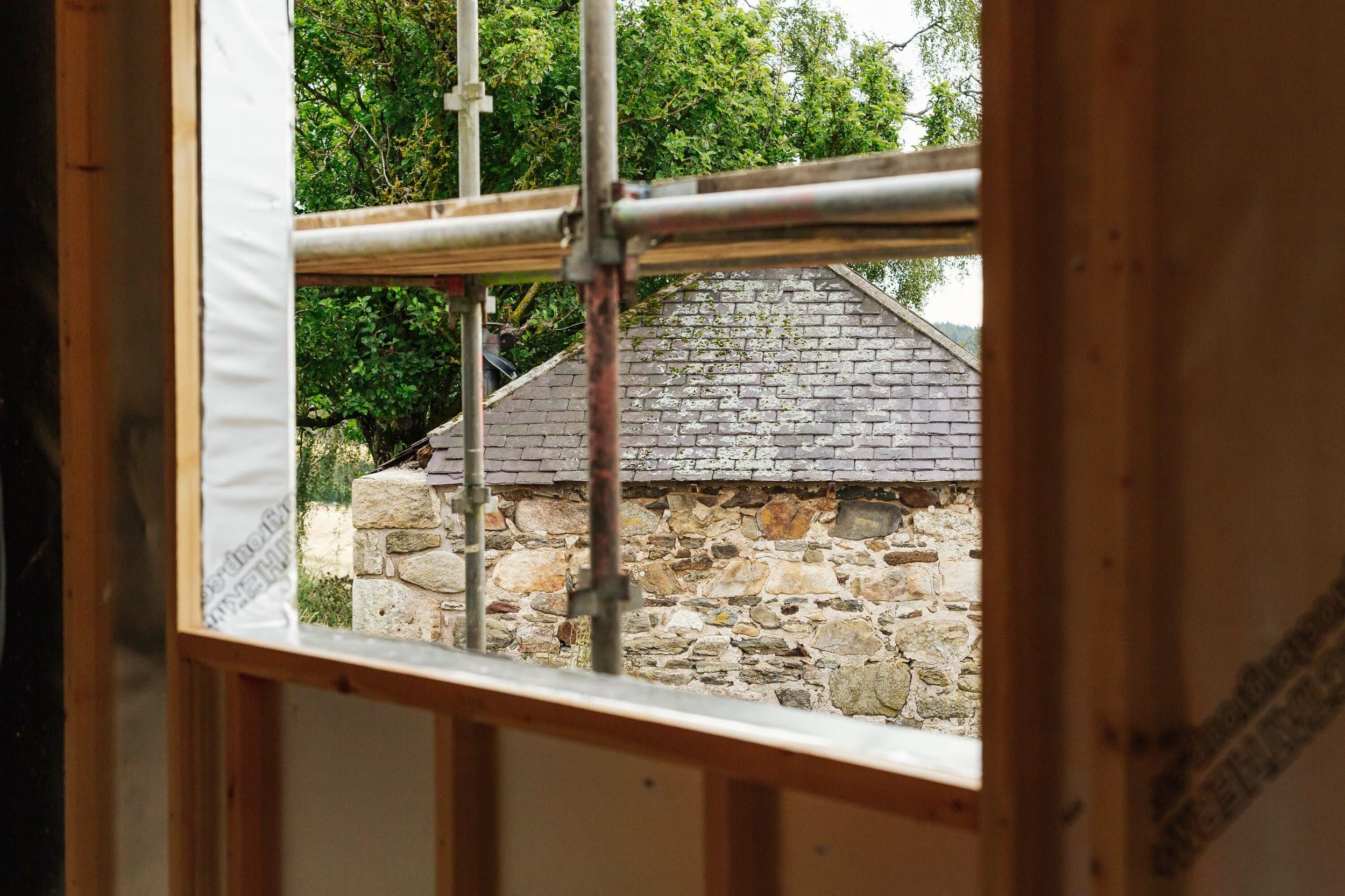 Looking through a window opening on a half-built house. A small stone croft, with a slate roof is in the background.