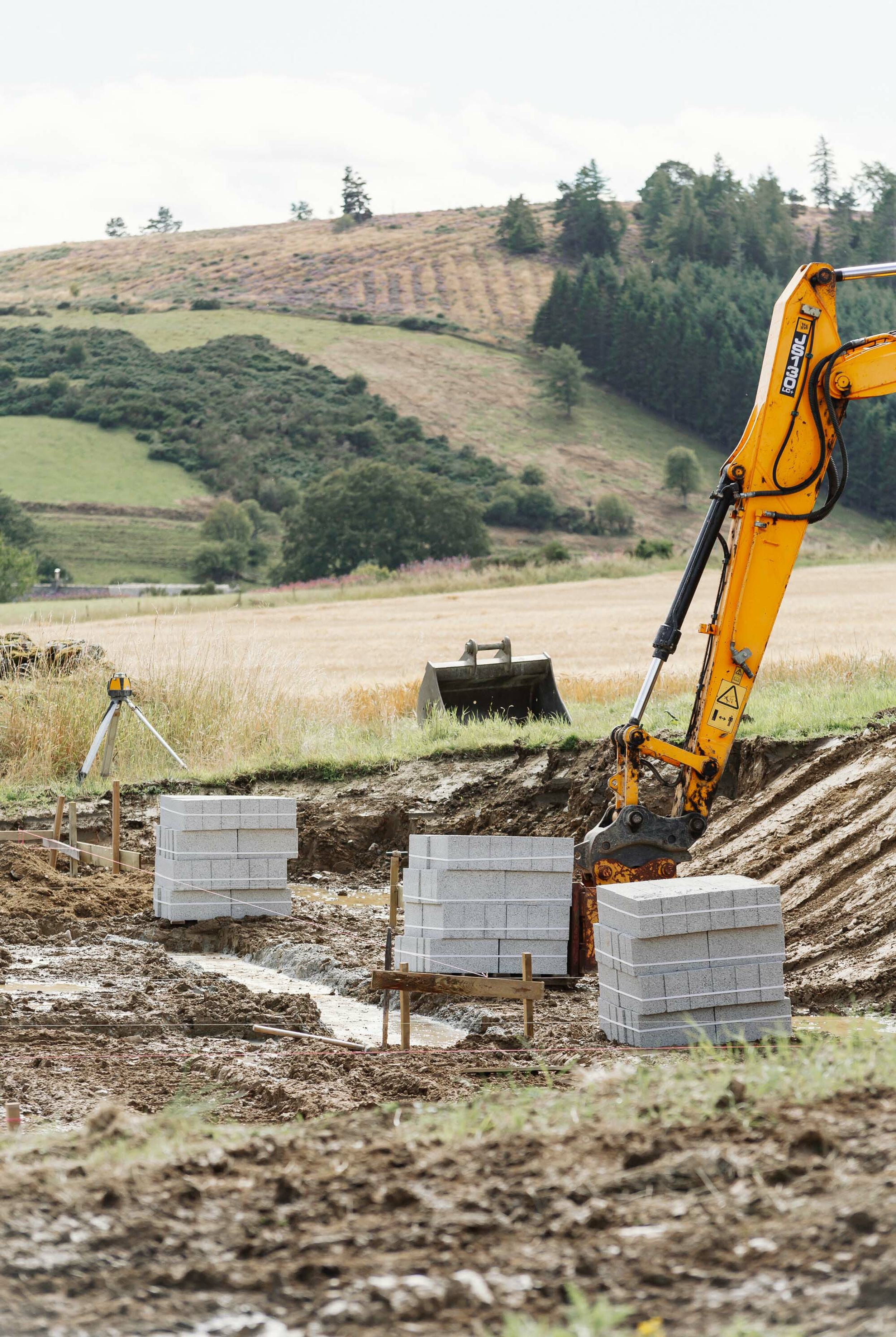 Stacks of grey concrete blocks sit next to the muddy foundations of new home. Builders line has been run around the permitter of the foundations. A yellow digger arm is visible. A digger bucket and laser level are in the background. A forest covered hill is in the distance.
