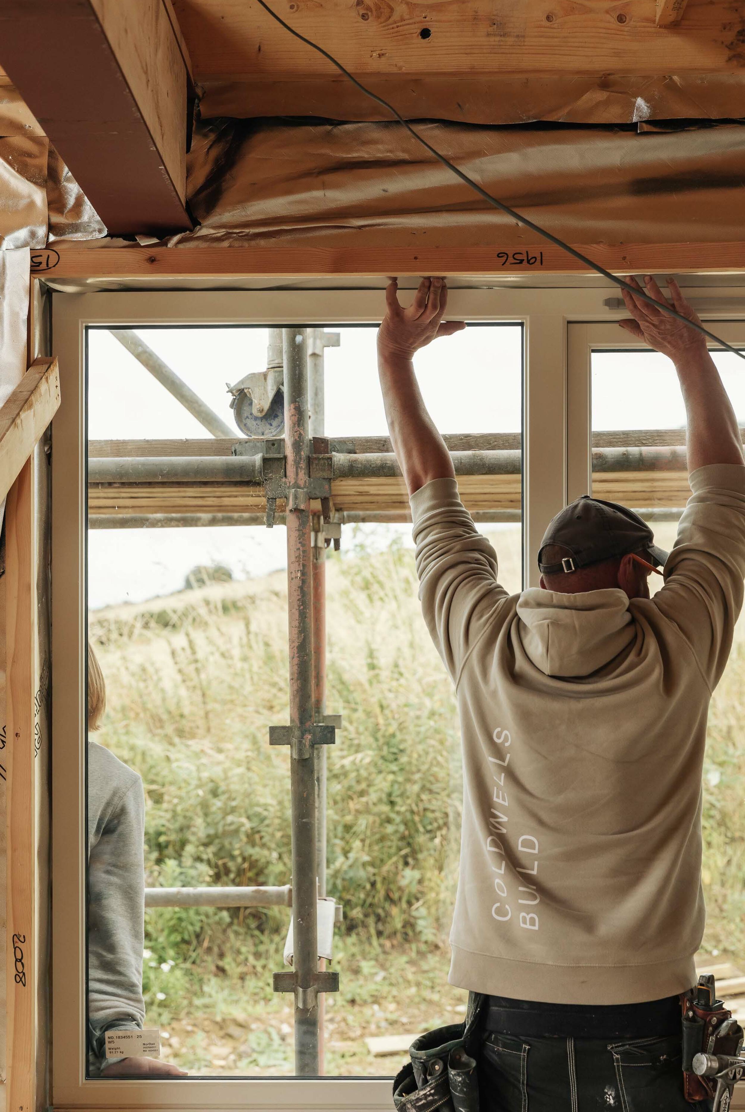 A builder stands inside a half-built house. He stretches up high to push a new window into its position. Outside, a builder supports the window.